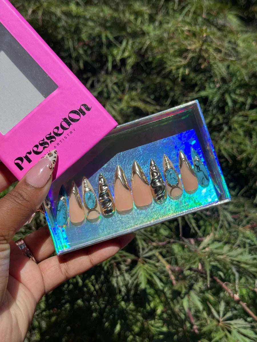 Medium-length press-on nails with a nude base and gold accents, featuring 3D turquoise decorations, snake embellishments, and a blue croc-like pattern on some nails.