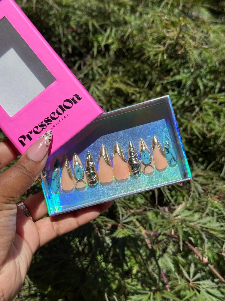 Medium-length press-on nails feature nude bases, gold accents, 3D silver snake embellishments, turquoise stones, and blue croc designs in almond and stiletto shapes.