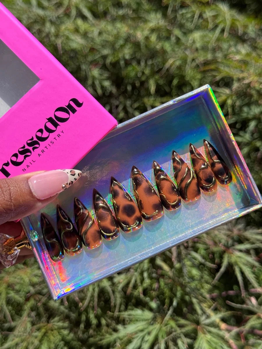 Medium stiletto press-on nails feature a brown cheetah print with gold accents, creating a bold, 3D design.