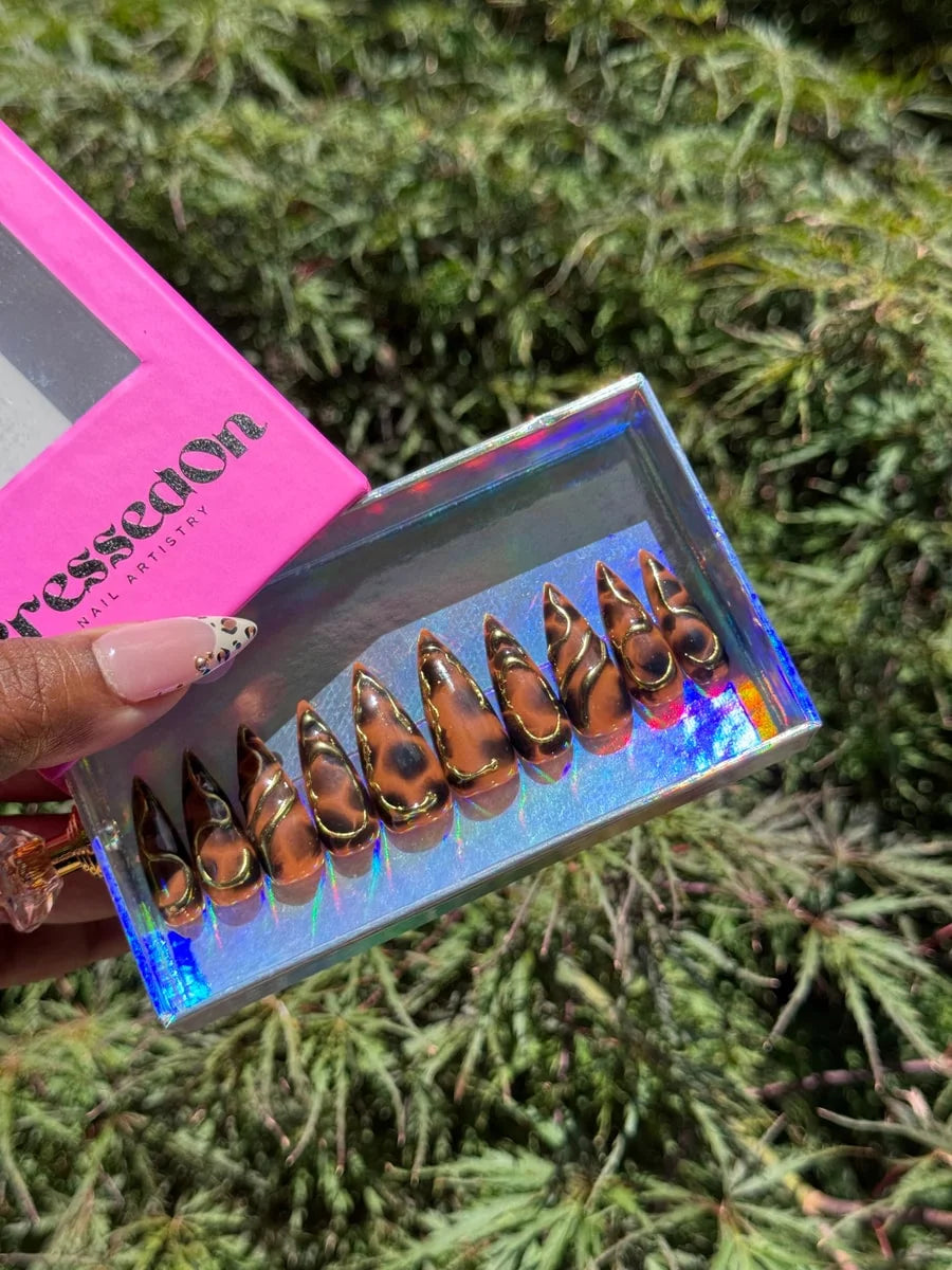 Medium stiletto press-on nails featuring a brown cheetah print with 3D metallic gold accents. The nails have a glossy finish.