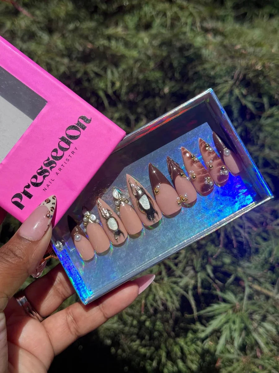 Medium length stiletto press-on nails featuring brown, nude, and leopard print designs. Nails have 3D embellishments, gold accents, pearl details, and abstract silver charms.