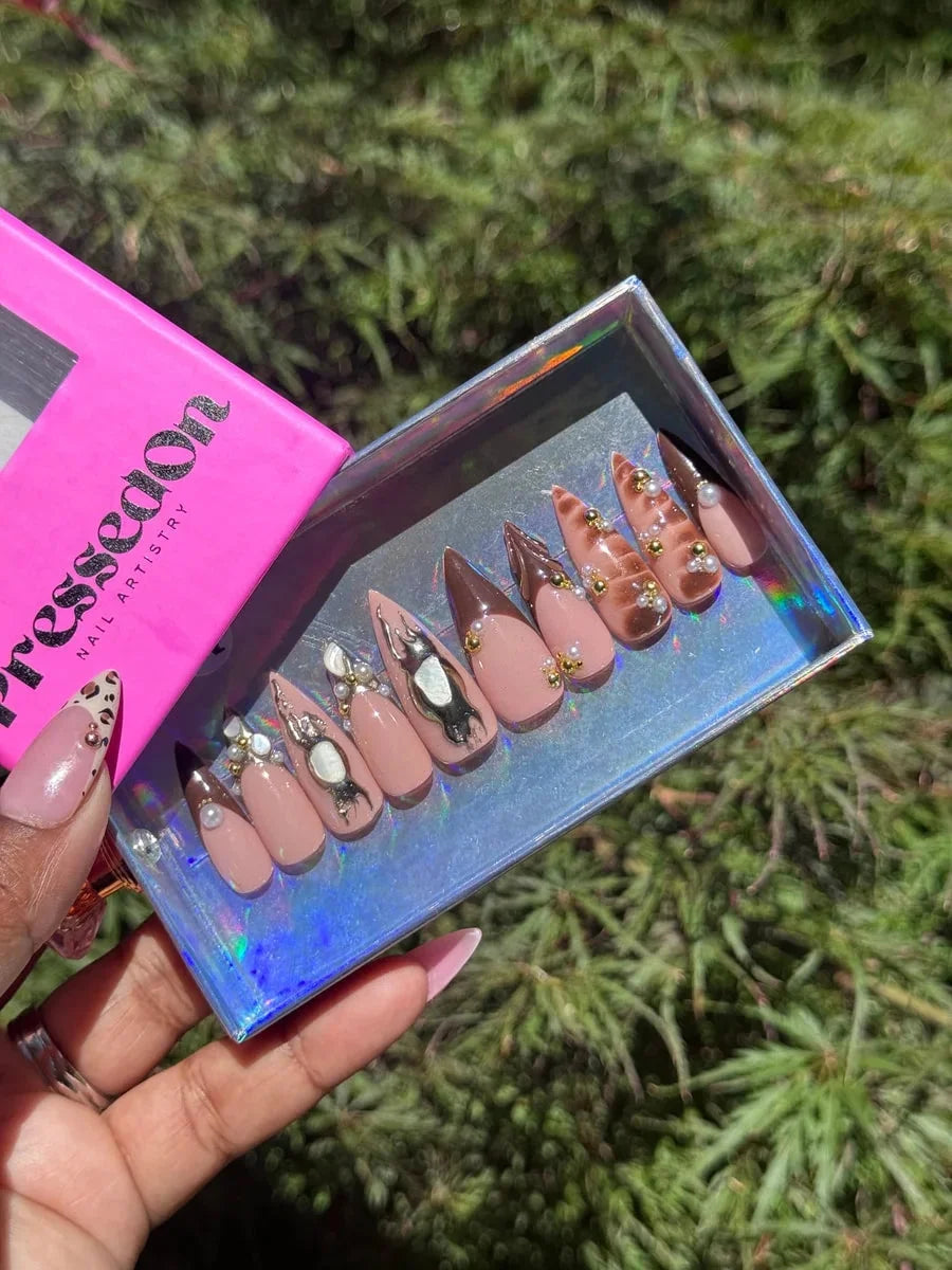 Set of medium length stiletto shaped press-on nails with varying brown and nude colors, featuring 3D decorations, gold details, pearls, abstract silver charms and crystal rhinestones.