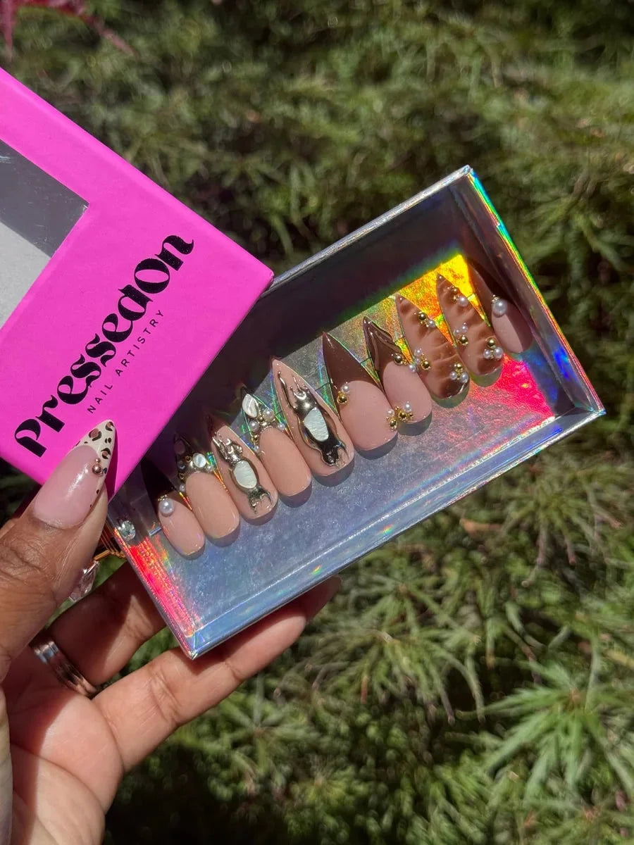 Medium length stiletto press-on nails feature brown, nude, and 3D accents, including brown tips, pearls, gold beads, crystal rhinestones and abstract silver charms.