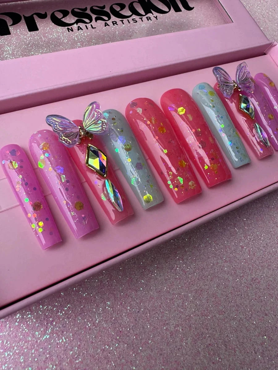 Long coffin press-on nails in pink and light blue with a glitter finish. The nails are decorated with 3D butterfly charms, rhinestone gems, and glitter flakes.