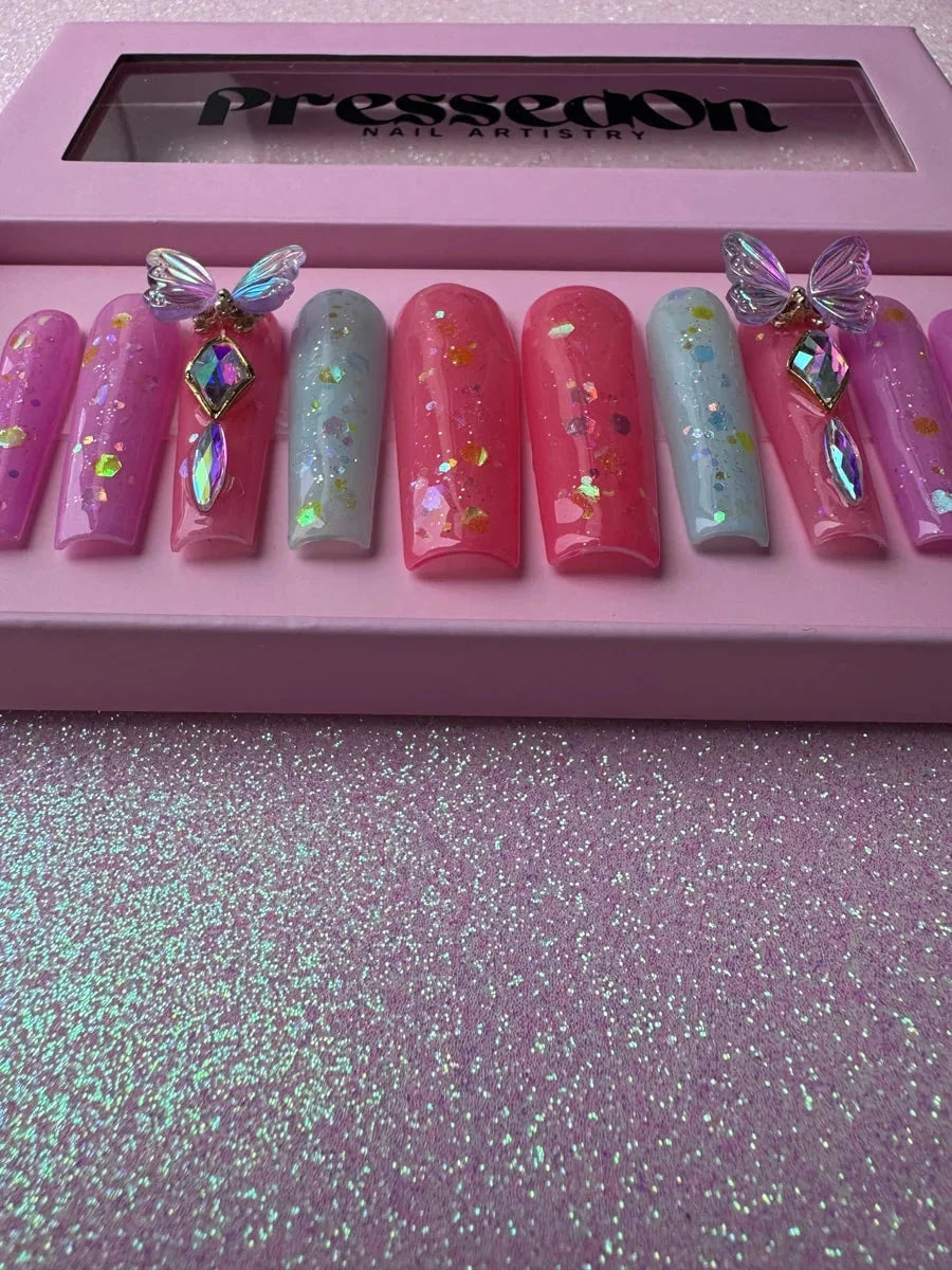 Long coffin press-on nails in pink, coral, and light blue with glitter. Two nails are decorated with 3D butterflies and rhinestone gems.