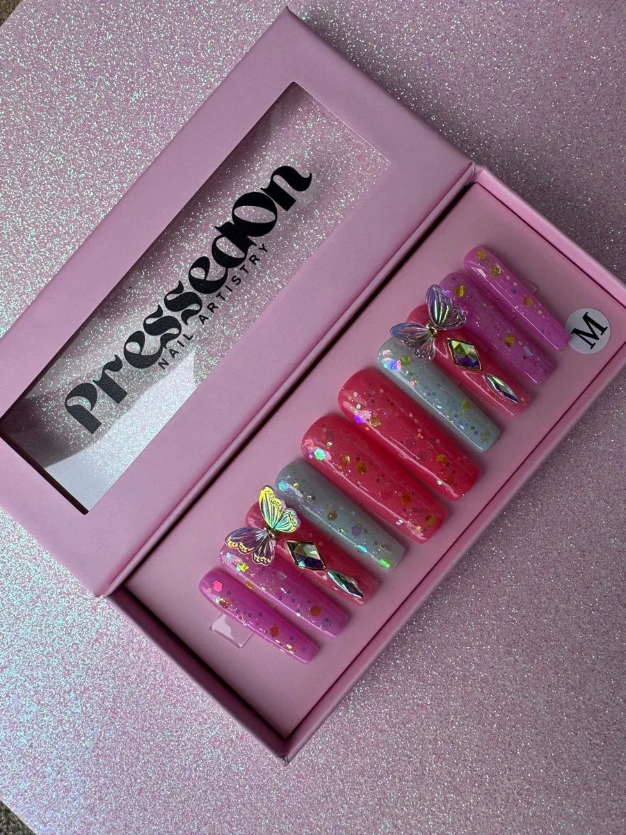 Long coffin press-on nails in shades of pink and blue with glitter accents, butterfly charms, and rhinestone gems. The set features 3D butterfly embellishments.