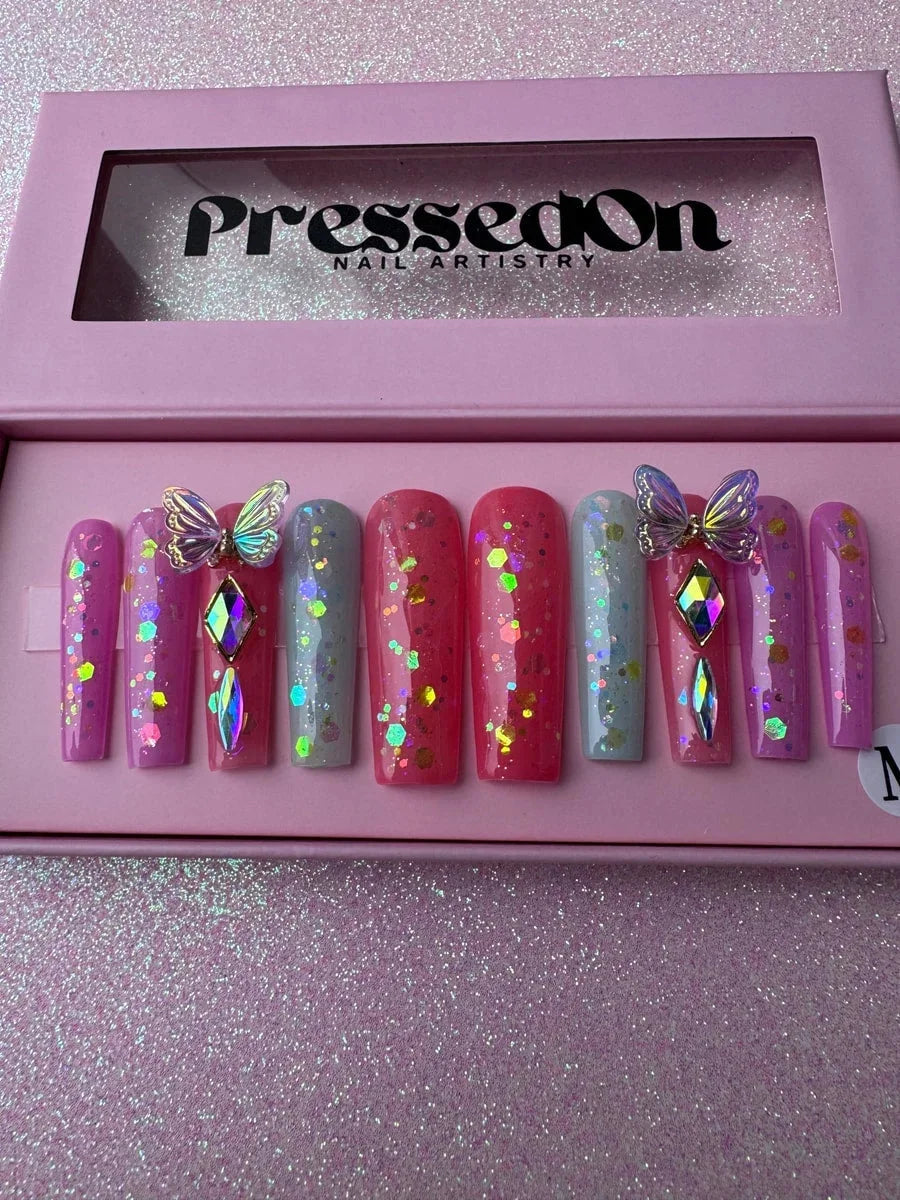 Long coffin press-on nails in shades of pink, coral and light blue with glitter and rhinestone gems. Two nails are decorated with iridescent 3D butterfly charms and gems.