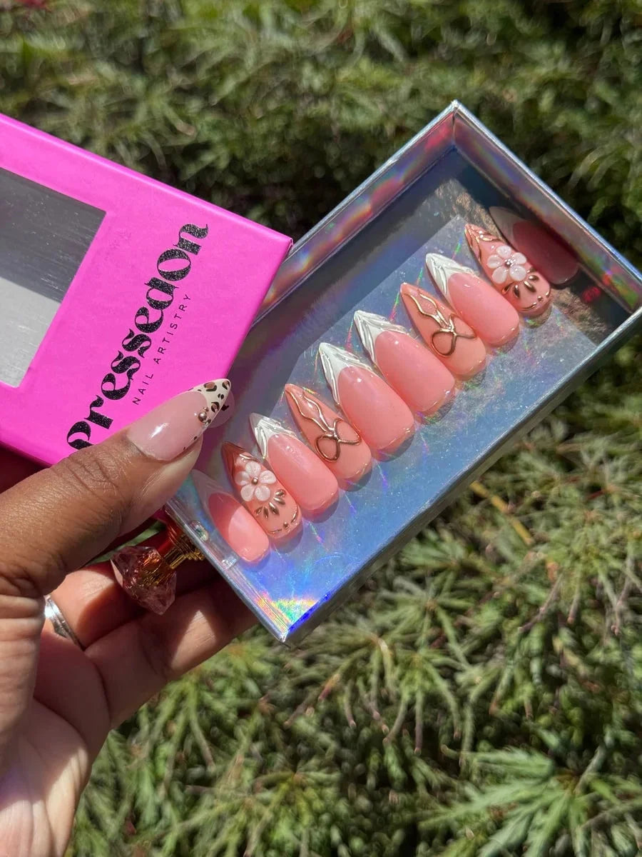 Set of medium pink press-on nails with white French tips, gold bow accents, and a 3D cherry blossom flower. One nail has a leopard print design.