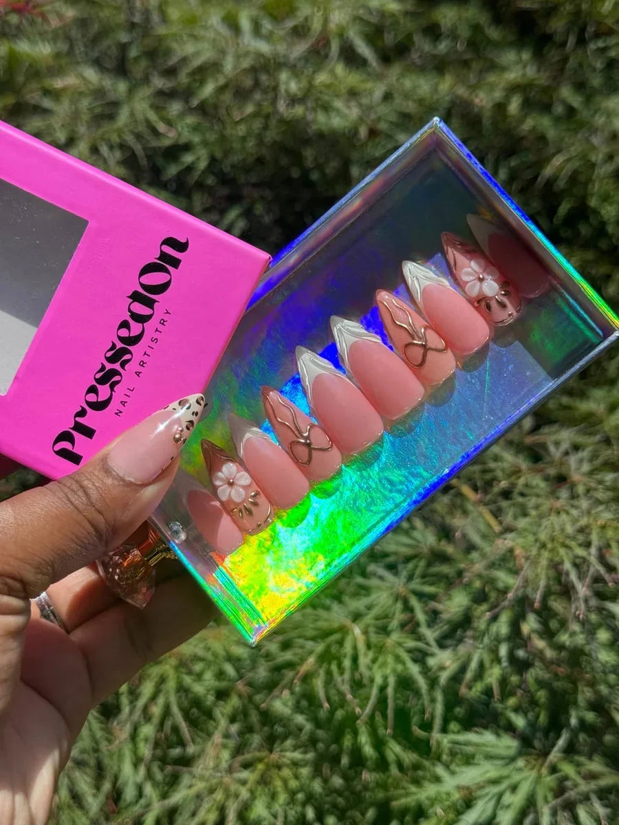 Medium-length pink press-on nails with white French tips are decorated with gold bows and a 3D cherry blossom flower. One nail has a leopard print design.