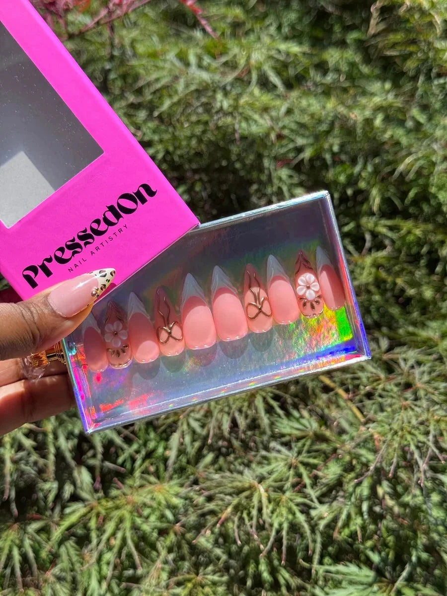 Medium pink press-on nails with white French tips, gold bow accents, and 3D cherry blossom flower embellishments.