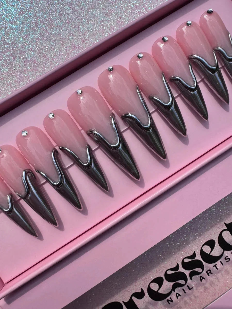 Long pink stiletto press-on nails feature a chrome 3D accent at the base and a small rhinestone near the cuticle. The nails have a glossy finish.