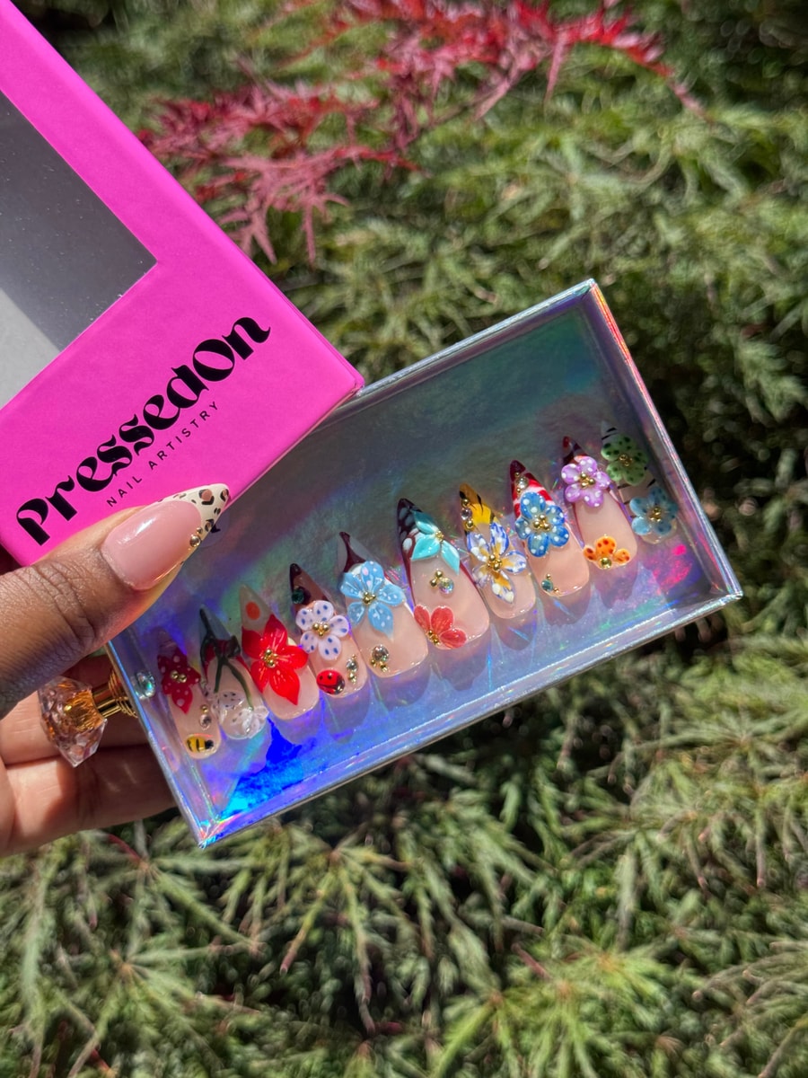 Nude press-on nails with 3D flowers in red, blue, purple, yellow, and orange colors, with rhinestone accents. A bee and ladybug are also featured.