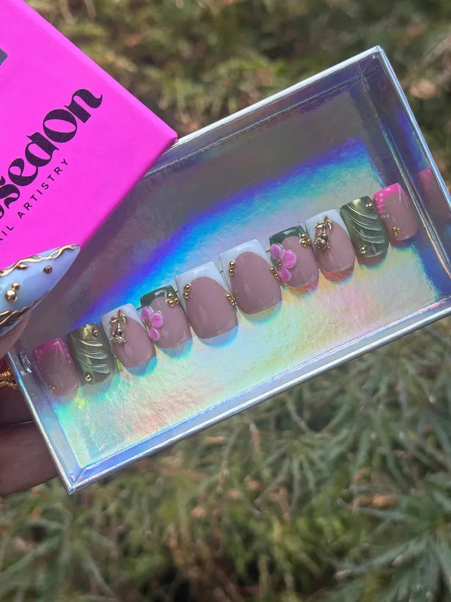 Short square press-on nails with a French tip design, featuring nude and white colors. Nails are decorated with 3D floral accents, gold beads, green wavy patterns, and pink tips.