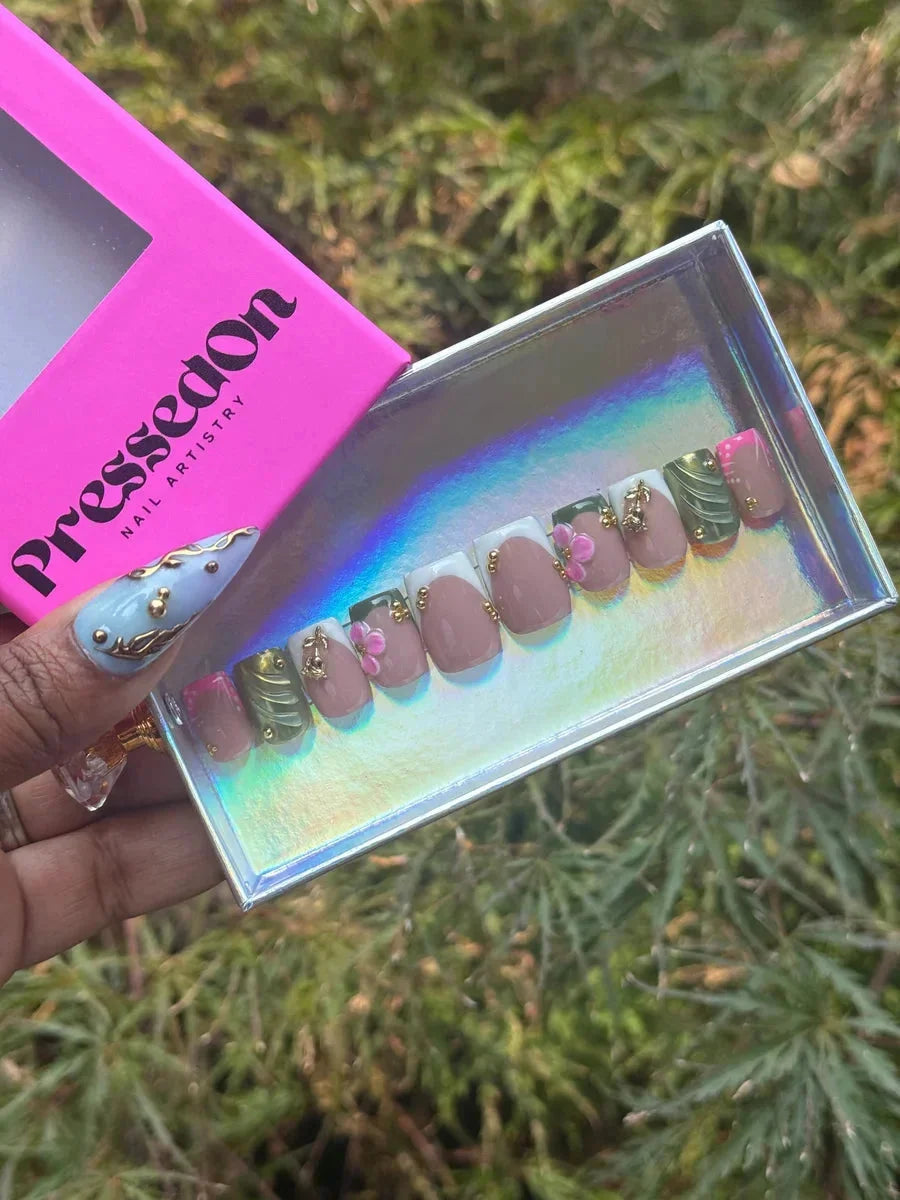 Short, square press-on nails with a French tip design, featuring 3D pink floral accents, gold embellishments, and green leaf patterns over a nude base.