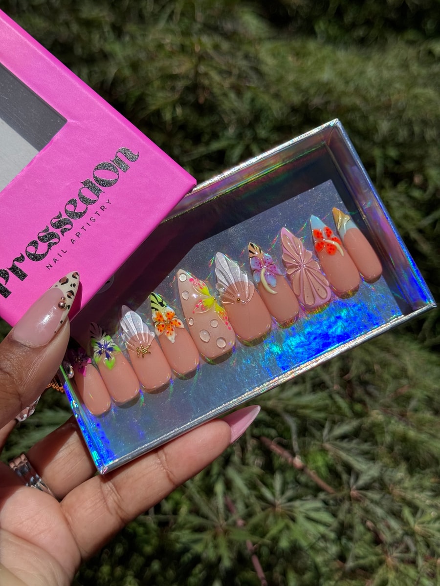 Medium-length nude press-on nails decorated with colorful 3D flowers, water droplets, and unique French tips. The design includes various floral accents and embellishments.