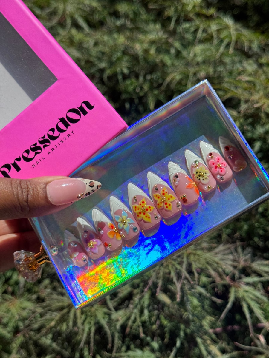 Short pink press-on nails featuring white French tips adorned with colorful 3D flowers, rhinestones, and gold embellishments.