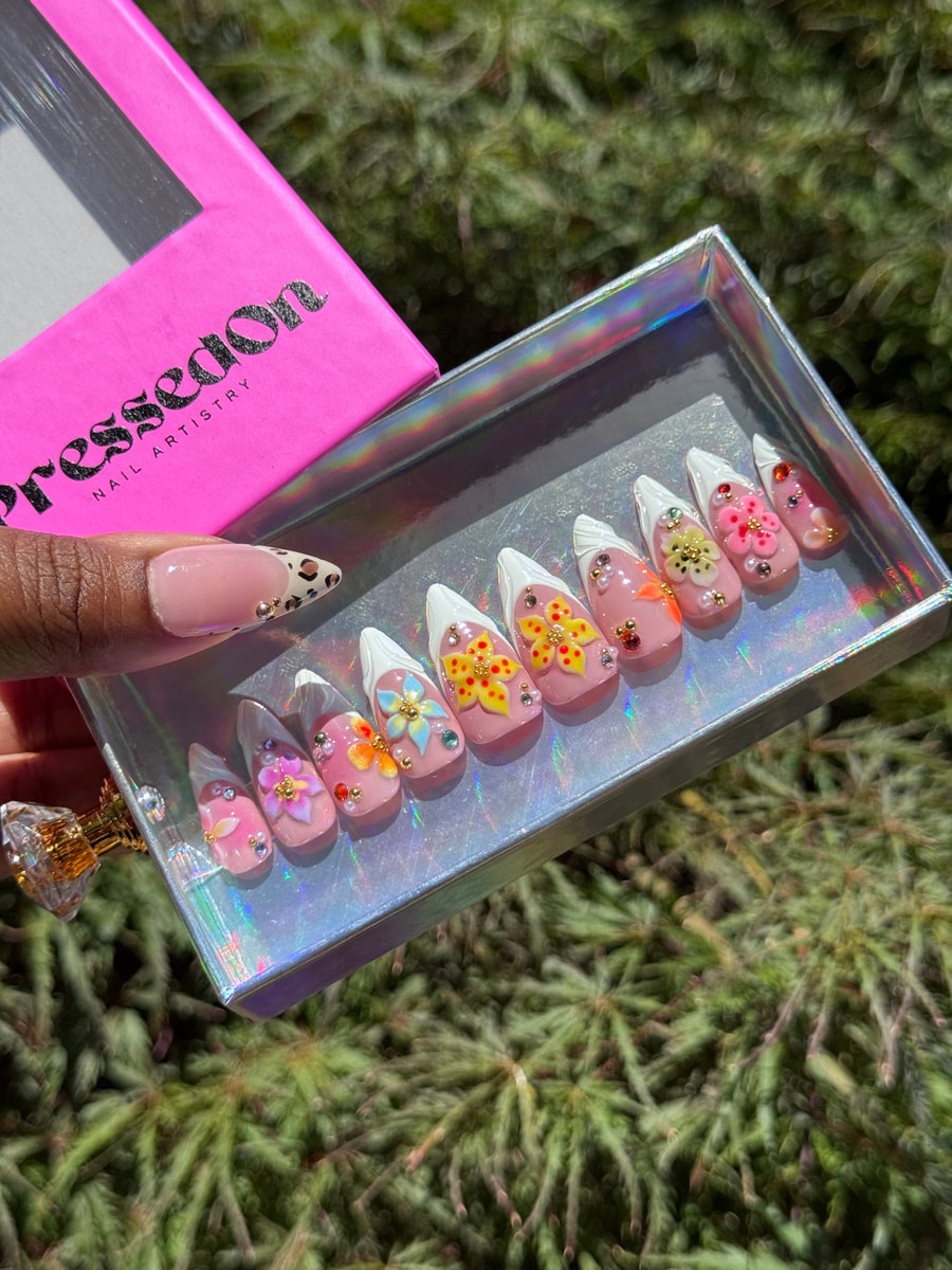 Short, pink press-on nails feature white French tips and colorful 3D flower accents, along with small gold beads and jewel embellishments.