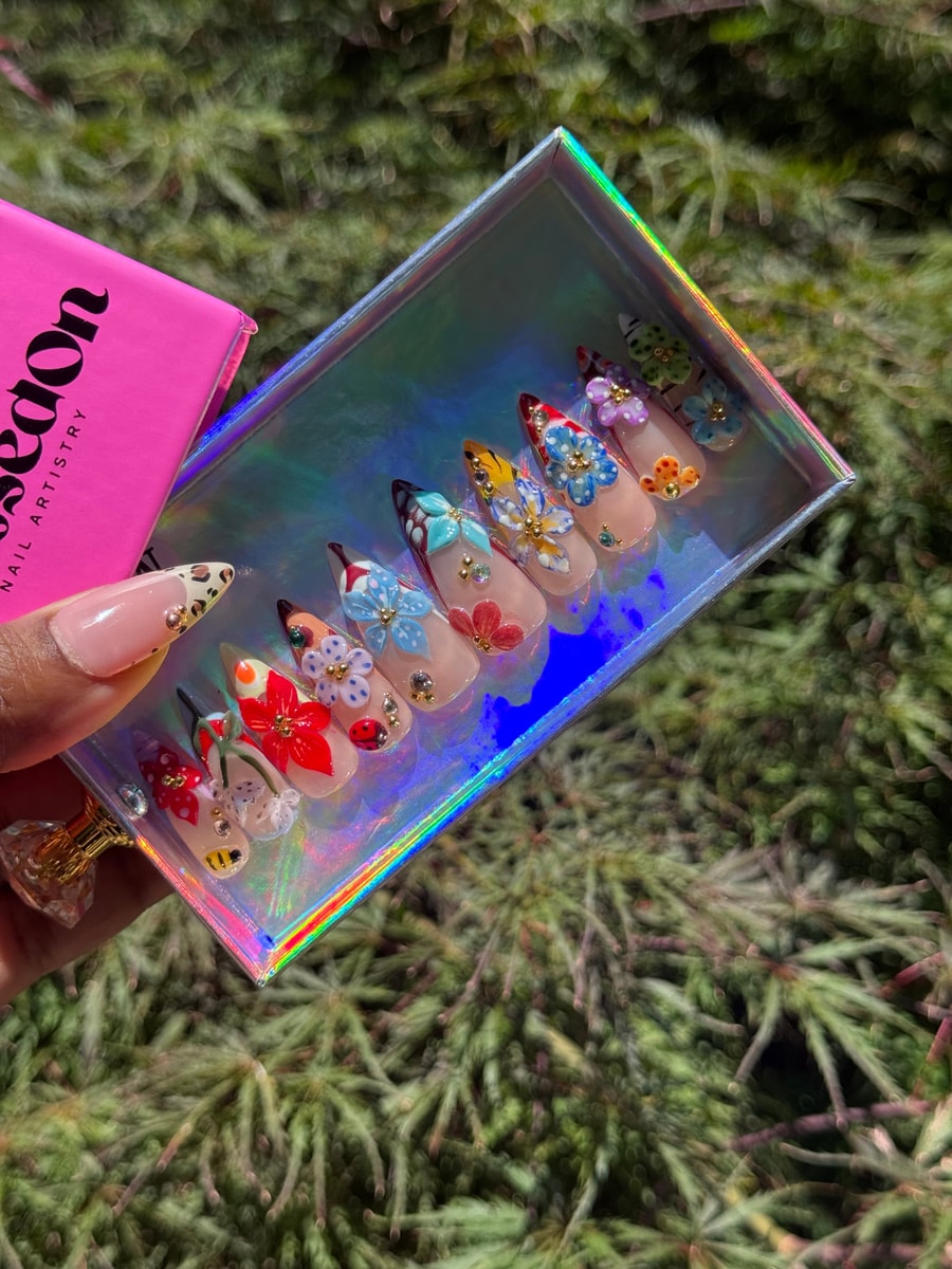 A set of medium length press-on nails decorated with 3D flowers in various colors, ladybug and bee details. The nails feature a mix of red, blue, purple, yellow, and green designs.