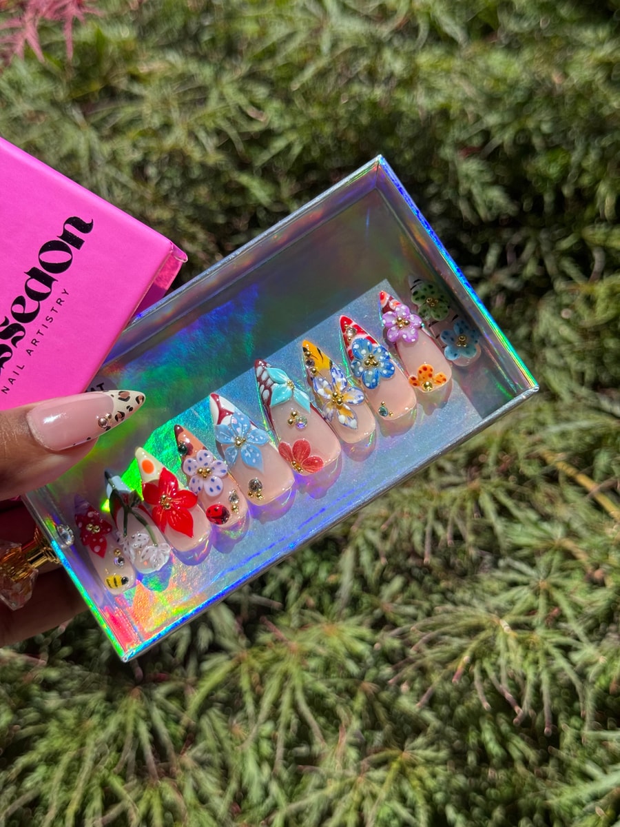A set of medium-length press-on nails with 3D flower and insect decorations. Designs include polka dots, butterflies, poinsettias, and a bumblebee, with gold accents and rhinestones.