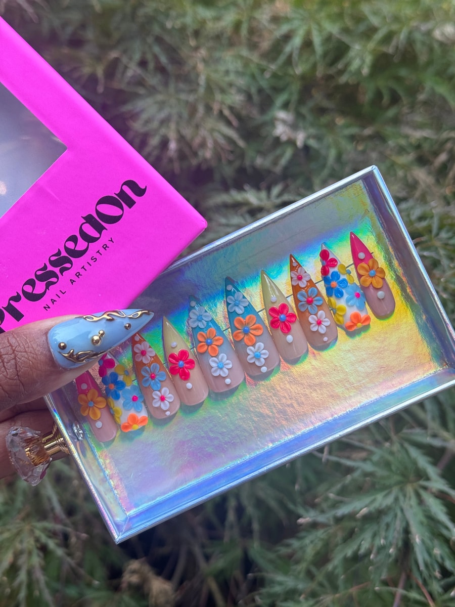 Short, stiletto press-on nails are decorated with 3D flowers in blue, orange, pink, and white. The base color is nude.