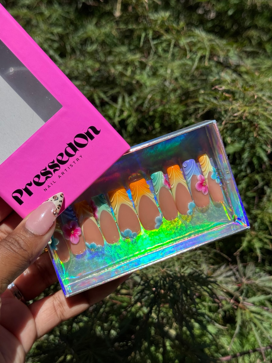 Medium length press-on nails featuring a brown base with colorful French tips and 3D flower accents. Various decorative rhinestones complete the look.