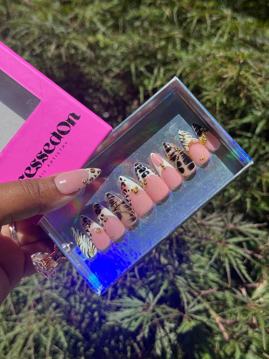 Short press-on nails feature animal prints like leopard and zebra, accented with gold details, on a pink base.