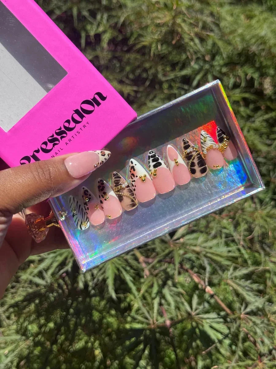 Short press-on nails with animal print designs, including leopard and zebra patterns, with gold accents. Some nails have a light pink base with animal print tips.