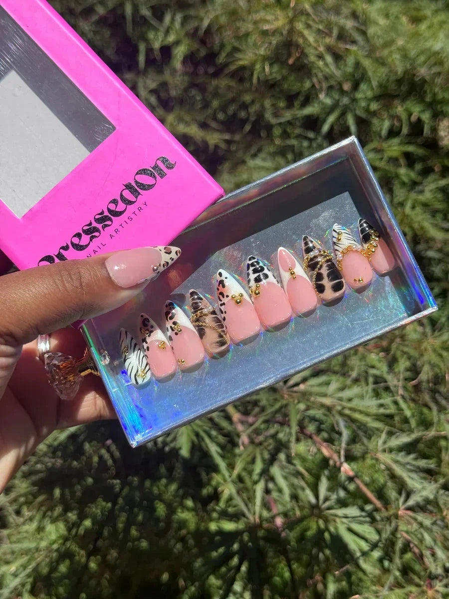 Short press-on nails with various animal print designs, including leopard and zebra patterns, with gold embellishments on a pink base.