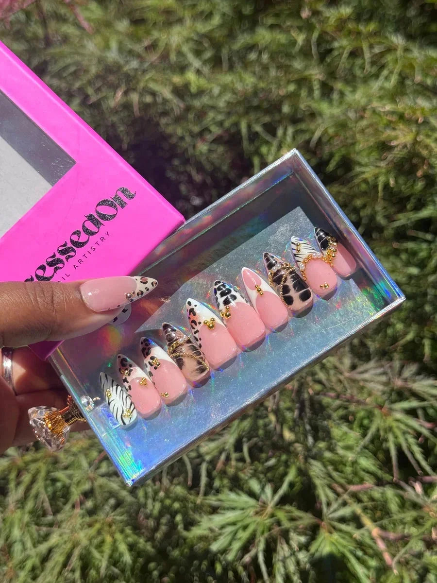 Short press-on nails feature a variety of animal prints, including leopard and zebra, with pink, gold, and black and white accents.