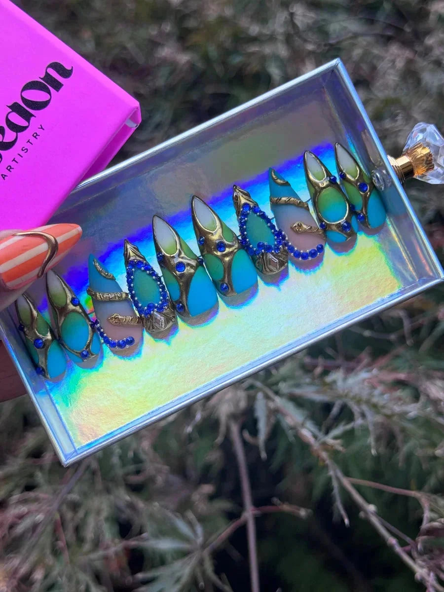Aqua pointed press-on nails feature blue gemstones, gold accents, and a unique snake design, with shades of aqua and seafoam green.