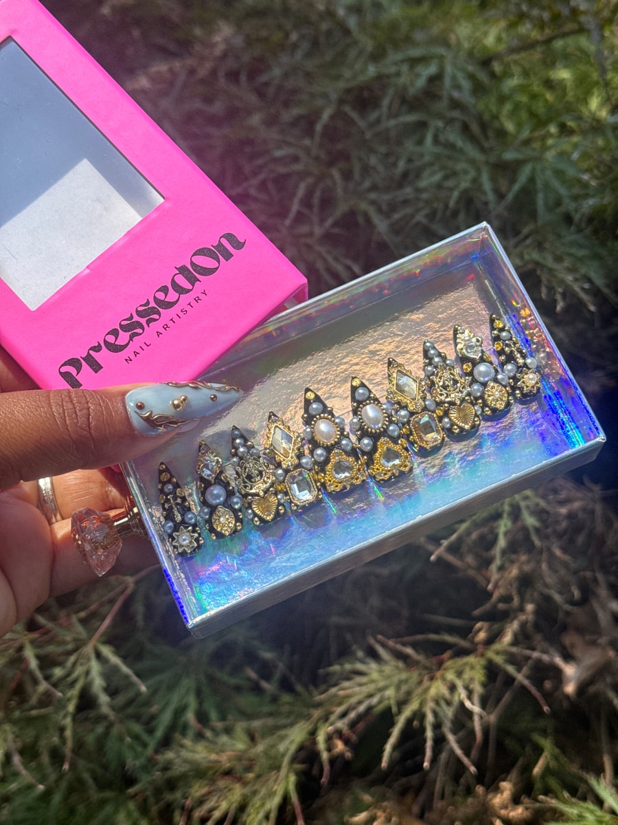 Short black press-on nails are decorated with a variety of gemstones, pearls, and gold accents. They include various shapes and sizes of stones and pearl embellishments.