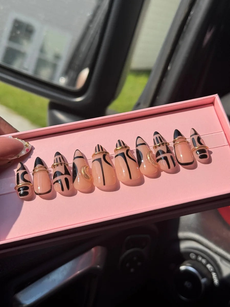 Set of short, nude-base press-on nails with black geometric designs. Some nails have small gold ring accents.