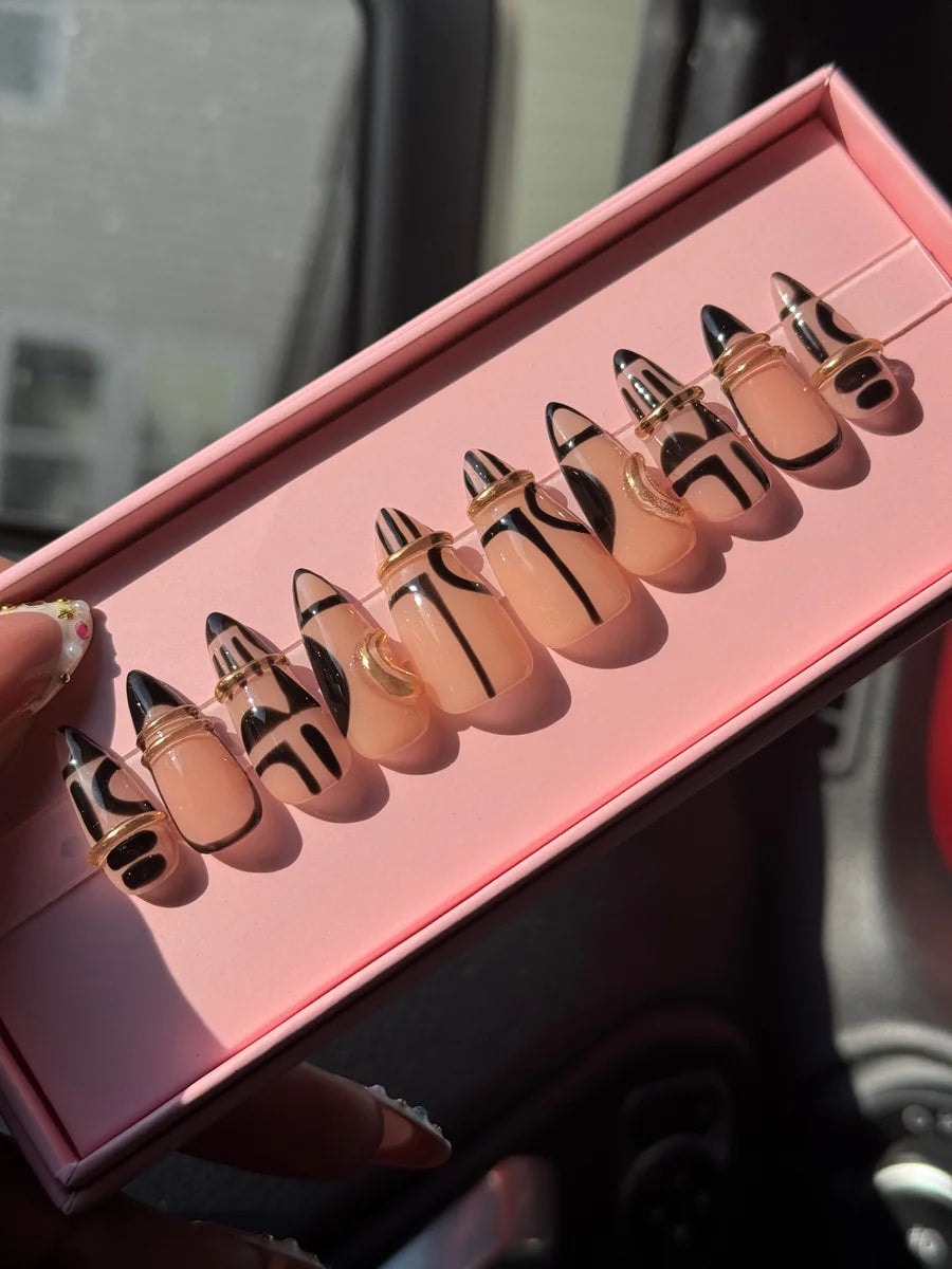 Short stiletto press-on nails with nude and black geometric patterns, each adorned with a gold ring.  The design features black accents and minimalist lines.