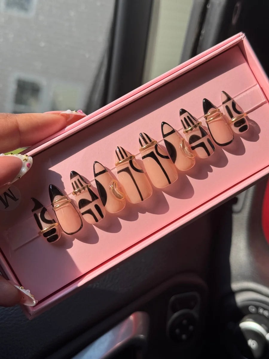 Short stiletto press-on nails feature a nude base with unique black geometric patterns and delicate gold wire accents on each nail.