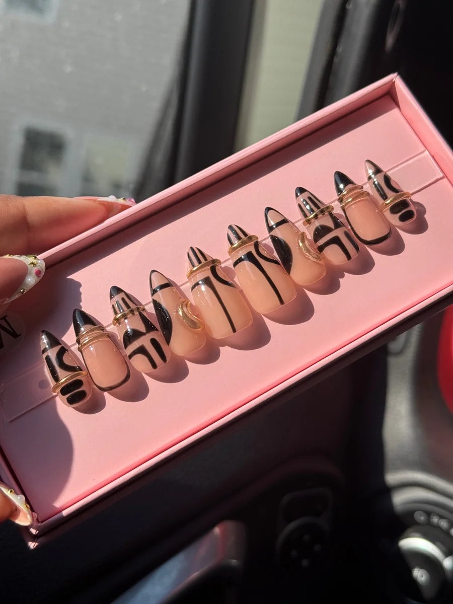 Short stiletto press-on nails with nude bases and black geometric line art. Each nail features a thin gold ring accent.