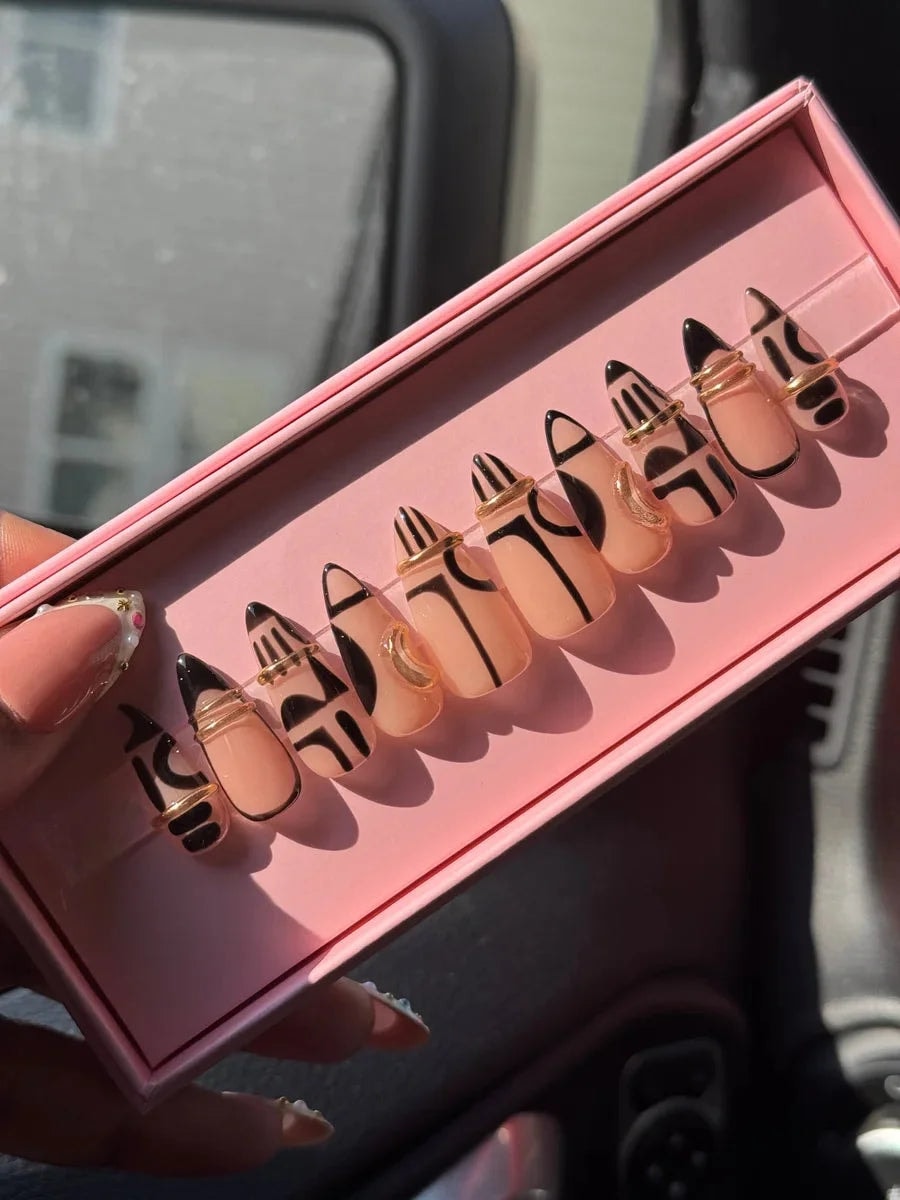 Short, nude press-on nails with black geometric patterns and thin gold wire accents.