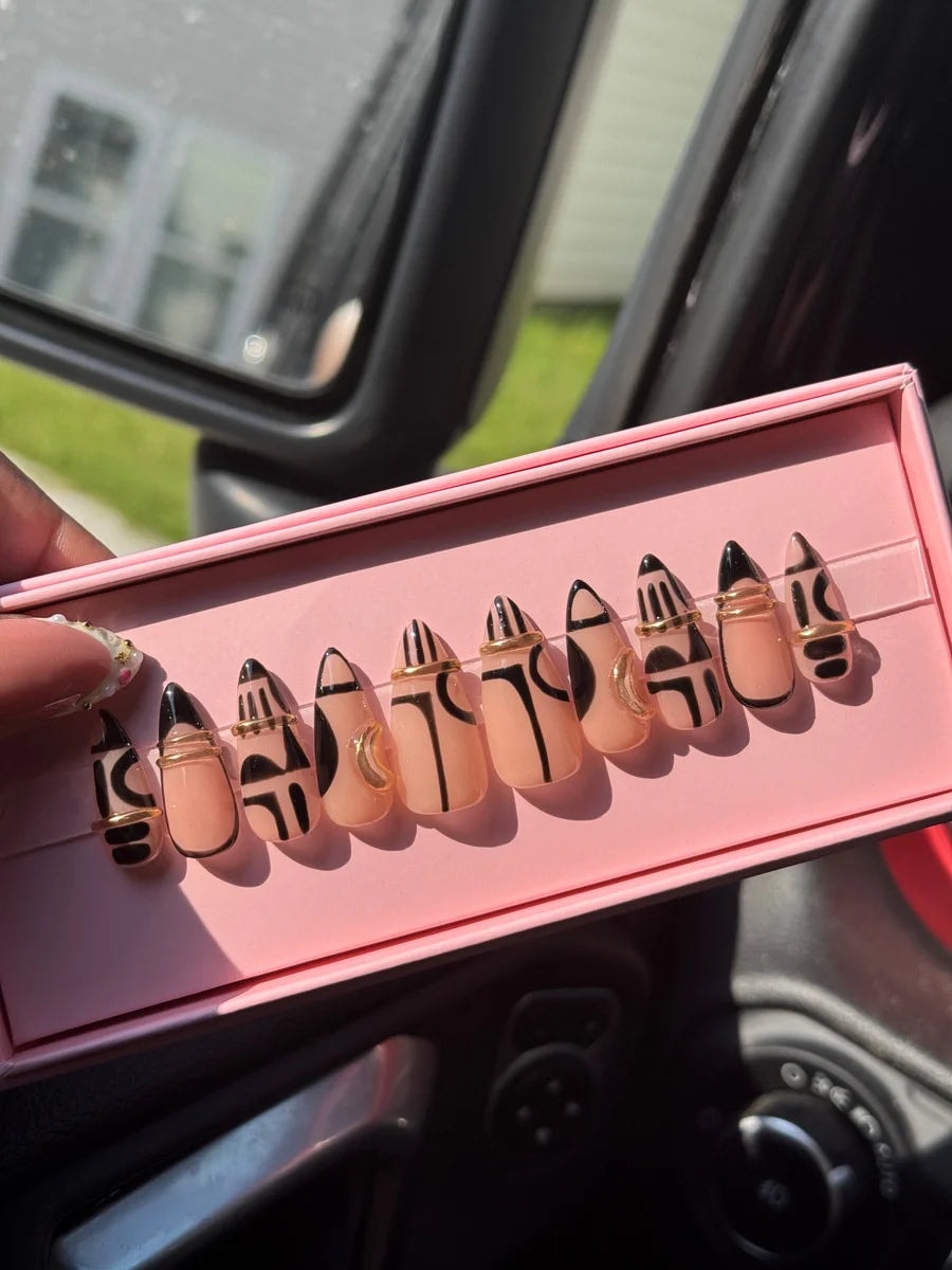These are short, almond-shaped press-on nails painted with a light pink base. Each nail has unique, bold black geometric designs and some nails have gold ring accents.