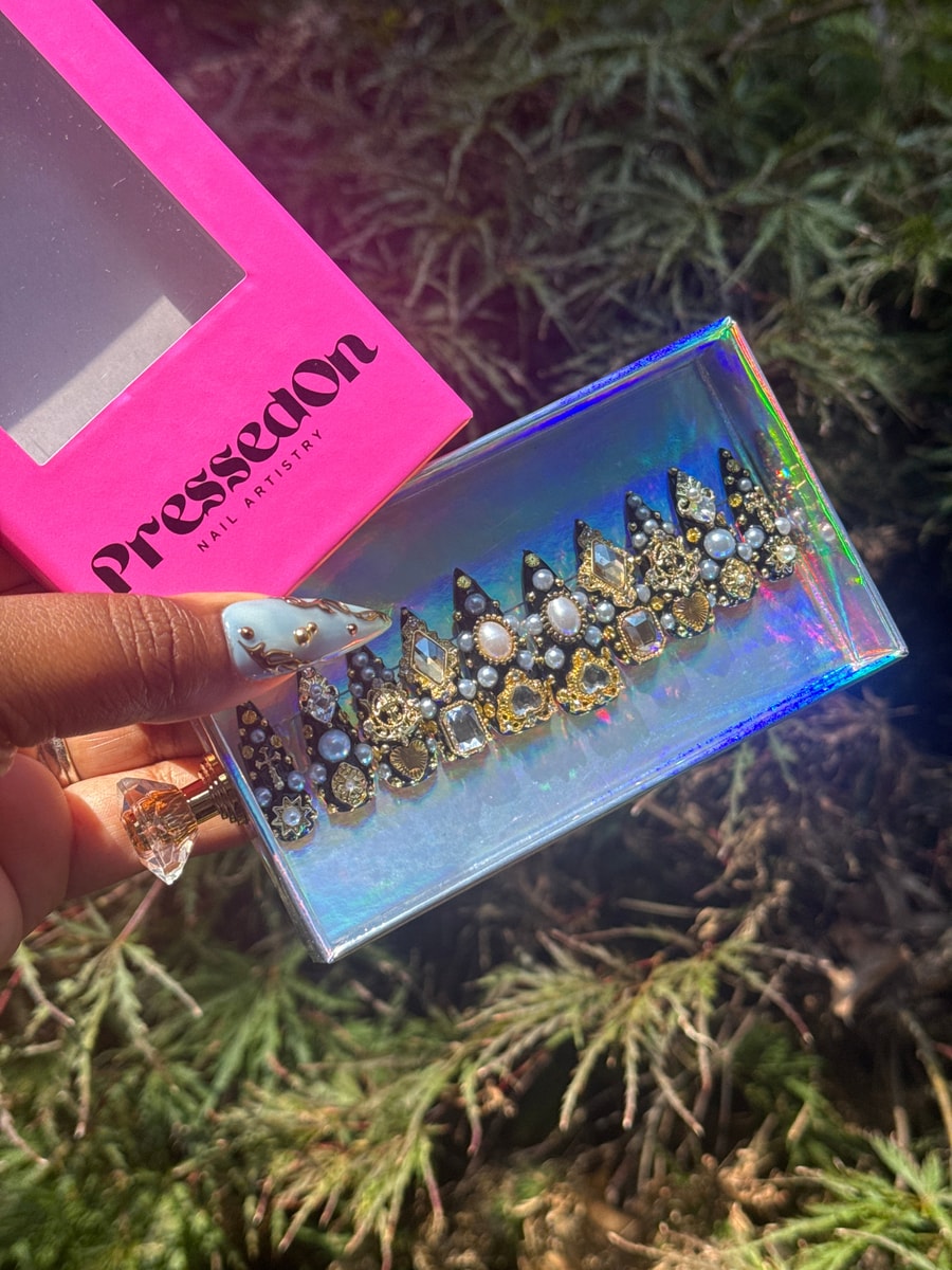 Short black press-on nails adorned with a variety of gems, pearls, and gold accents in assorted shapes and sizes.