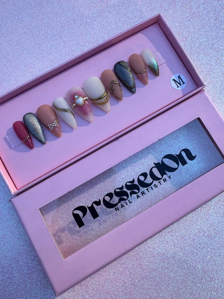 A set of medium almond-shaped press-on nails featuring pink, mauve, silver glitter, and solid color designs, accented with gold chains, rhinestones, and pearl embellishments.