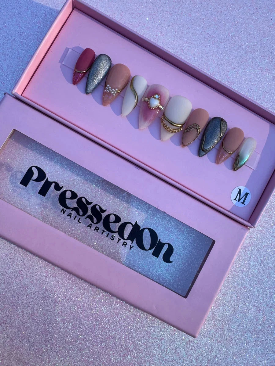 A set of medium almond press-on nails with bling freestyle designs. Features glitter, solid colors, gold chains, pearl embellishments, rhinestone accents and gem details in pink, gray, nude and white shades.