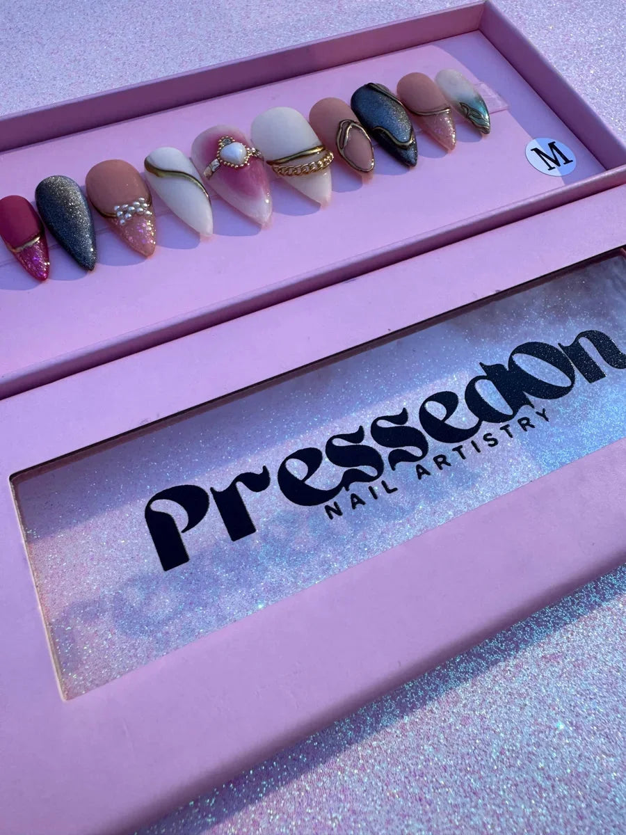 Medium almond-shaped press-on nails featuring pink, nude, gray, and white designs with glitter and bling. Details include gold chains, pearl beads, rhinestones, and gem embellishments.