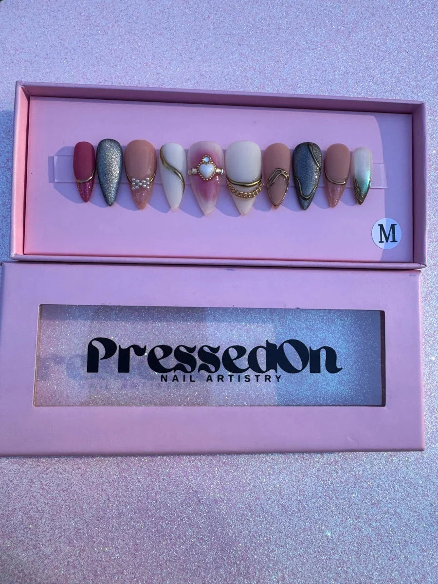 Set of medium almond-shaped press-on nails with glitter, pink, silver, and nude shades with gold accents, pearl embellishments, heart charms and rhinestone accents.