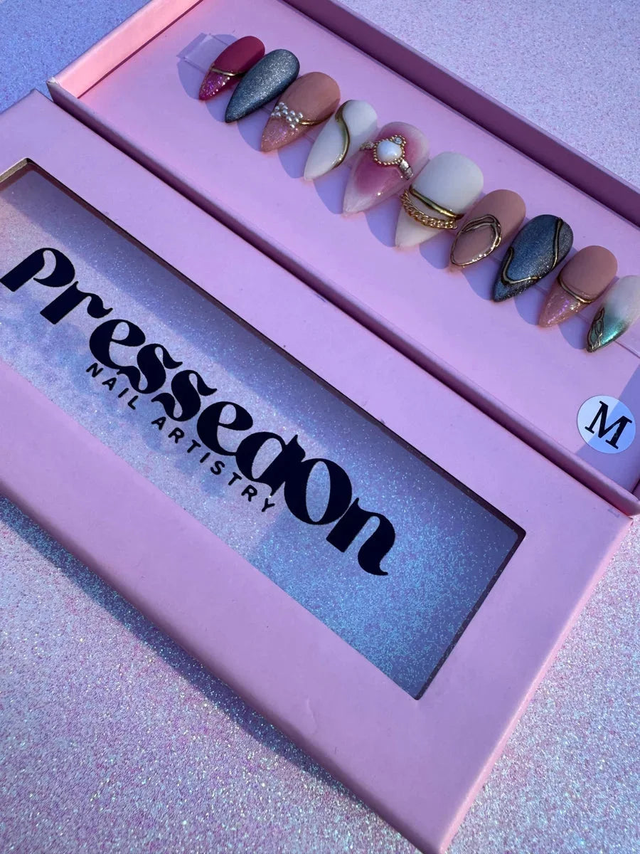 Medium almond-shaped press-on nails featuring freestyle designs with pink, silver glitter, and nude shades. Bling accents include gold chains, pearl embellishments, and gem details.