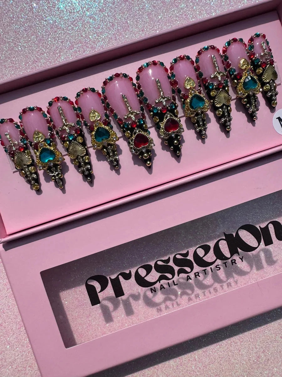 Set of medium stiletto press-on nails featuring a pink base, black tips, and colorful rhinestones and gems. The nails are embellished with gold hearts, crosses, and bead accents.