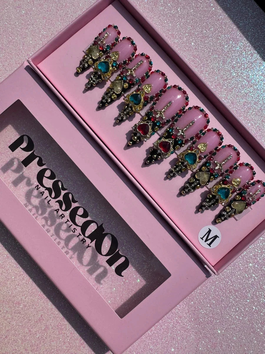 Medium pink stiletto press-on nails with multicolored jewels, gold hearts and crosses, and black tips. The blinged-out design features rhinestones and bead accents.