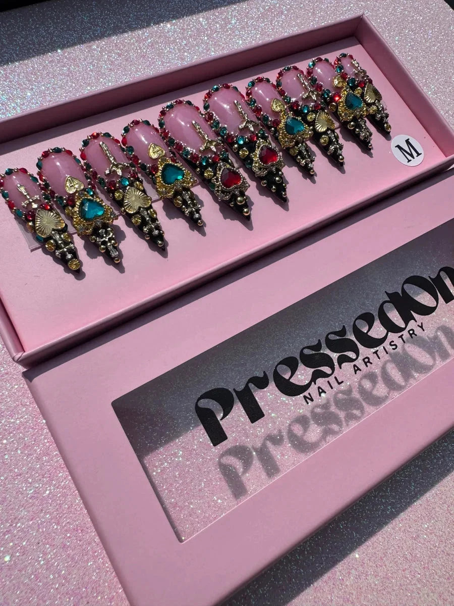 Medium stiletto press-on nails with a pink base, embellished with jewels, rhinestones, gold hearts and crosses, and black tips. A blinged-out design.