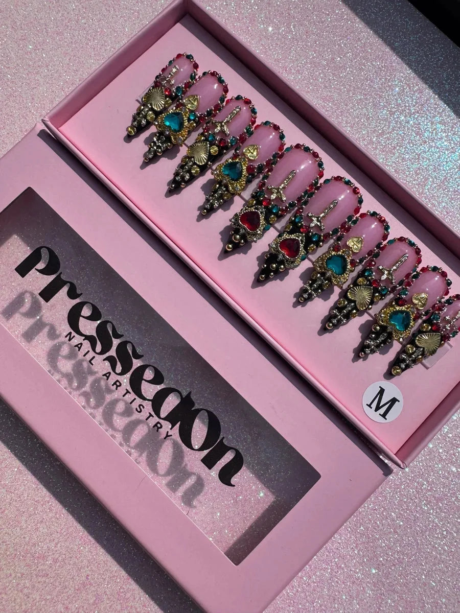 Medium stiletto press-on nails feature a pink base with ornate red, teal, and gold jewels, heart and cross charms, rhinestones and black tips with gold accents.