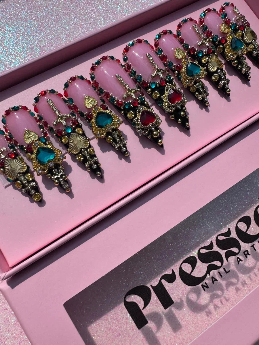 Medium length pink stiletto press-on nails are decorated with colorful jewels, red and teal gems, gold Hearts and Crosses, Rhinestones, and black accents on the tips.