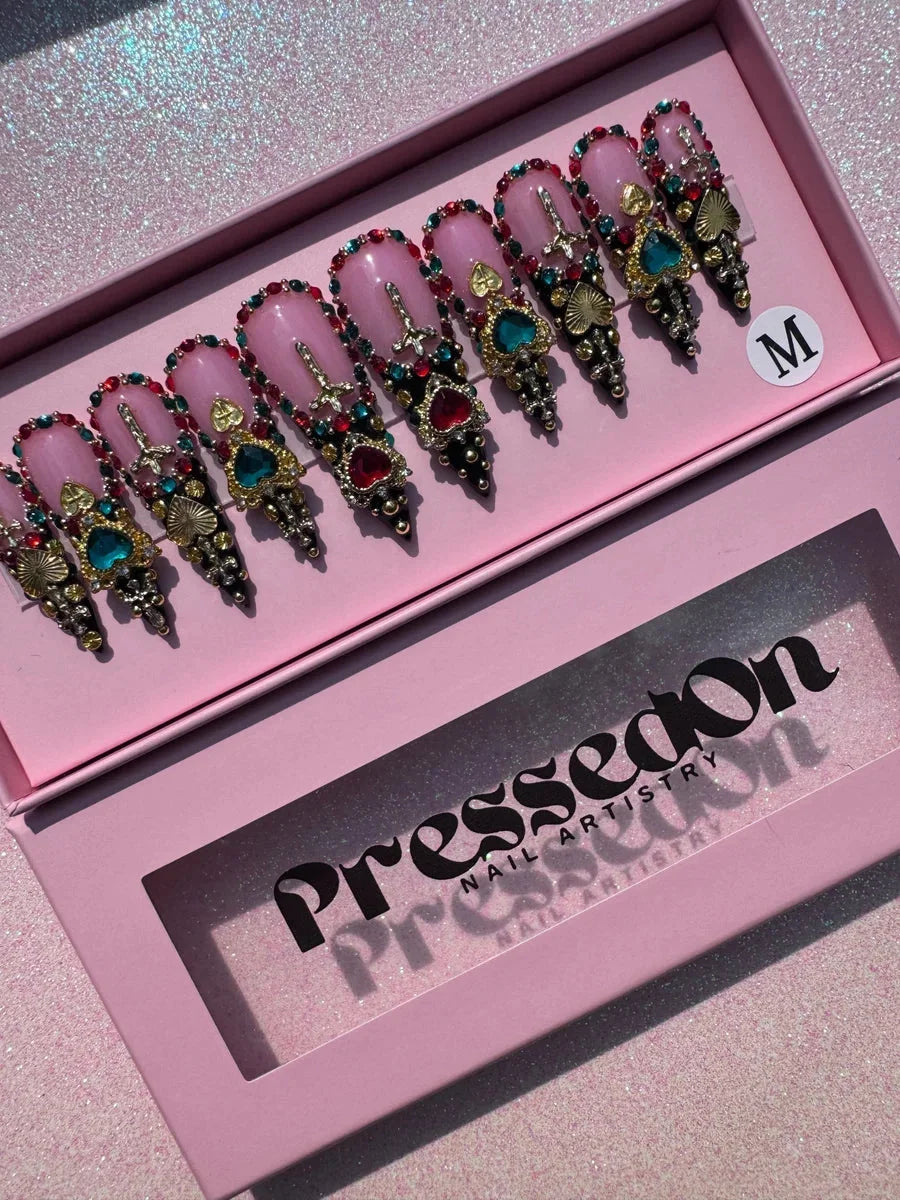 These are medium stiletto press-on nails with a pink base. They are heavily decorated with colorful jewels, hearts, crosses, rhinestones, gold embellishments, and black accents.