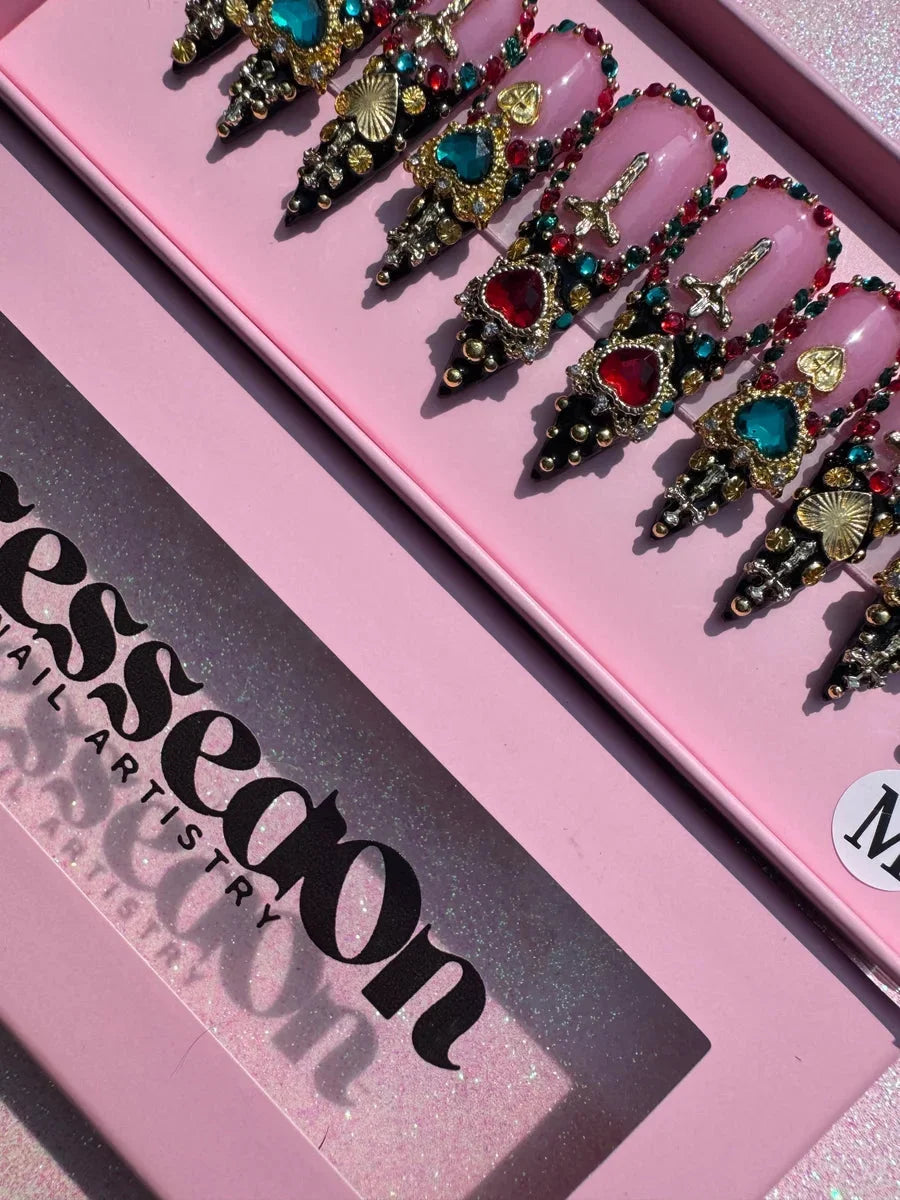 Medium-length stiletto press-on nails featuring a pink base, multi-colored jewels, gold heart and cross charms, rhinestones, and black pointed tips with gold accents.